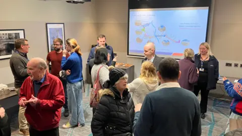 Wide of drop-in event shopping twelve people chatting in small groups in front of an information screen