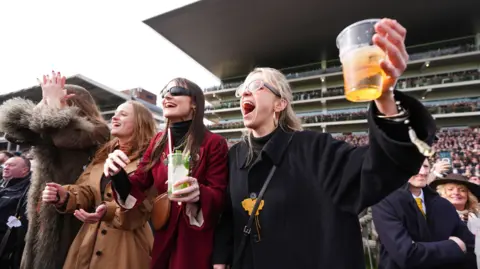 A row of four women stand in the cheering in the stands at Cheltenham Racecourse. They wear heavy coats and sunglasses. 
