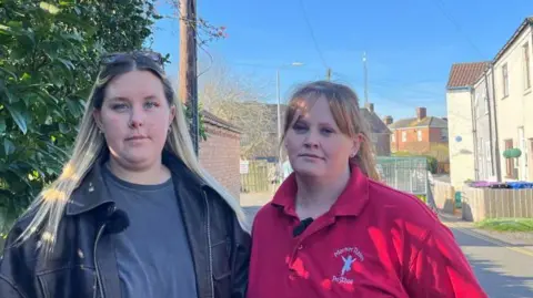 Two women look into the camera as they stand by the side of a narrow road. The woman on the left has dark-blond hair and wears a black leather jacket, grey-blue T-shirt and sunglasses on the top of her head. The woman on the right has ginger hair, tied back, and wears a red polo shirt. Green shrubs and white-washed houses can be seen. The sky is blue.