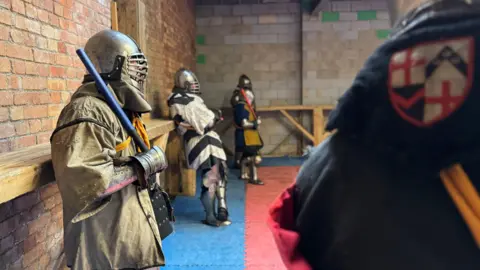 Men in armour and helmets lean against a wooden railing before fighting each other.