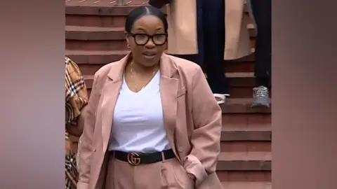 A woman pictured walking. She is wearing a light brown suit and white t-shirt. She has a slick black middle part hairstyle.
