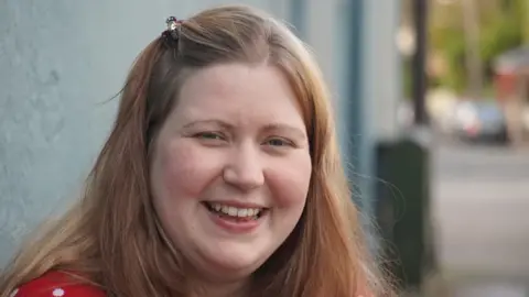 BBC/Alleycats TV A woman smiles at the camera with long brown hair, clipped back off her face. She wears a red top. There is a street behind her.