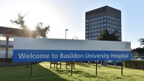 An exterior view of a hospital building. In the foreground is a sign which reads "Welcome to Basildon University Hospital". In the background is a tall grey tower block.