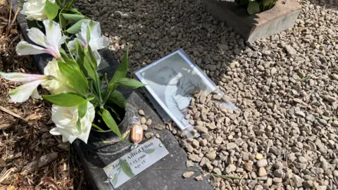 Nathan Turvey/BBC A picture of James Alfred Elton, one of the children buried in the grave, next to a plaque with his name on it with cream flowers in a pot