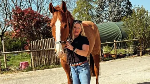 Kayleigh Sweet A blonde white woman with black sunglasses over her head wearing a black shirt and blue jeans is standing next to and holding a brown horse with a white patch on its nose.