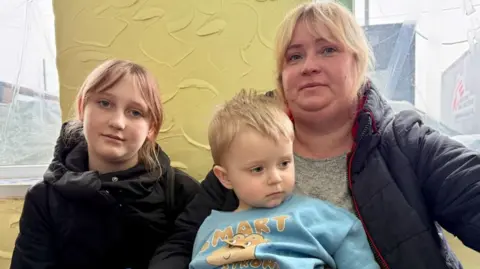 Matthew Goddard/BBC Kateryna, a woman with blonde hair wearing a quilted coat, poses for a photo with her children - a young girl of about 11 and a small boy of about four