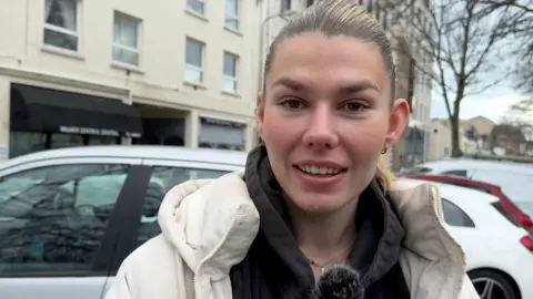 Imogen Brooks standing in a car park. She has brown tied back hair and a white coat.