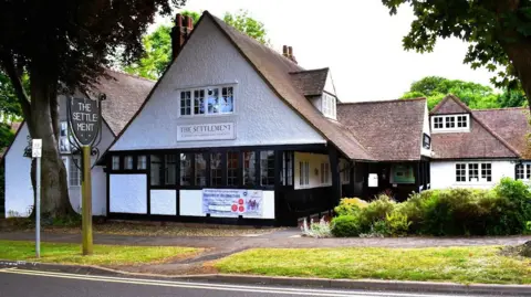 Historic England A two storey early 20th Century building in Letchworth Garden City. It has steeply pitched tiled roofs, with dormer windows and the walls are white render with black frames to wide windows. There is a pavement and grass verge in front and a sign on a post saying The Settlement in block capitals. 