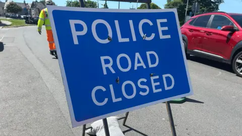 A road sign which reads: 'POLICE. ROAD CLOSED.' There is a man wearing high-vis behind the sign and a red car off to the right hand side.