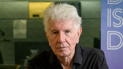 BBC A man with white hair stares at the camera, wearing a black cord shirt, in a studio