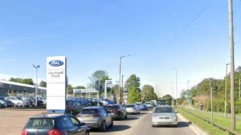 Redbourn Road, Hemel Hempstead, with cars on the road, by a car dealership. There are trees and grass to the left, and signs for the dealership to the right. 