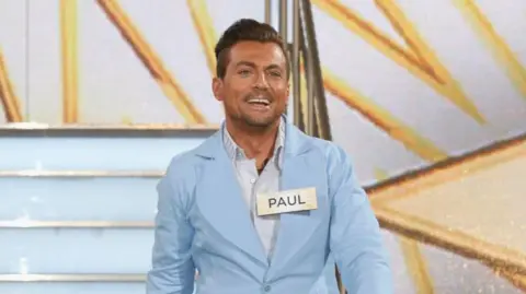 Getty Images Paul Danan wearing a pale blue suit and a large gold name tag. He is walking onto stage at the premiere of Celebrity Big Brother. He has blue eyes and dark hair which is swept backwards and shaven short on the sides. He looks happy and tanned. 