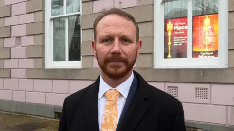 Tom Coles is stood in front of the States Assembly building. He is wearing a black jacket and yellow tie
