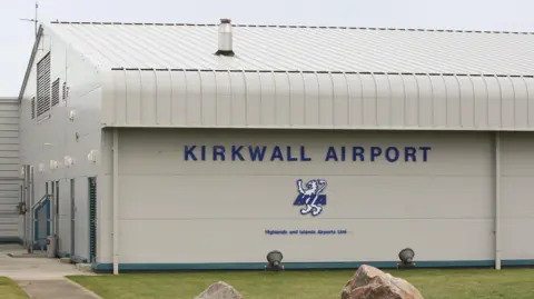 A general view of Kirkwall Airport. The terminal building is grey, with the name and a lion motif in blue and white on the side. There is a patch of green grass in front and the sky above is grey.