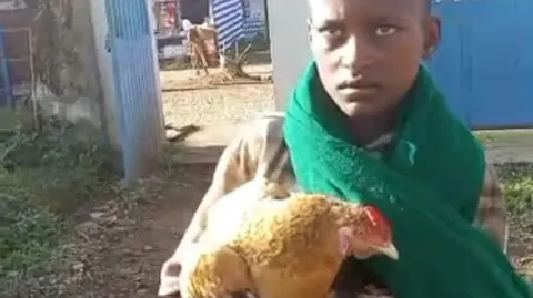 Markos Abaye wears a green scarf and holds a brown chicken