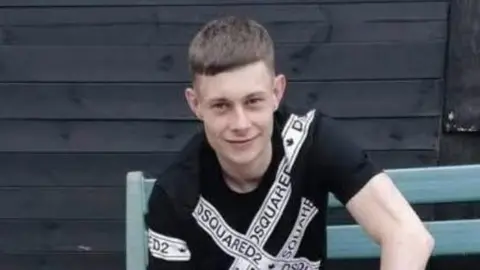 A young man with short brown hair wearing a back t shirt with white bands across it and the text Squared2. He is sat on a blue wooden bench with a shed behind him and is smiling at the camera. It is a head and shoulders shot of him.