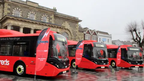 Three red electric buses lined up in front of buildings in Salisbury. There are red flags next to them.