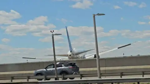 A plane flying over a bridge which a car is driving on. The plane is just past the the bridge and it is very low in the air 