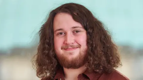Joe Wrieden smiles at the camera. He has shoulder length thick curly brown hair and a gingery beard.