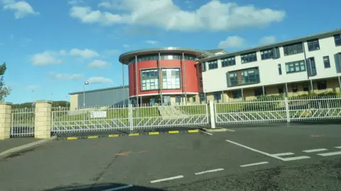 A striking secondary school building with a round, red section prominent behind a set of school gates