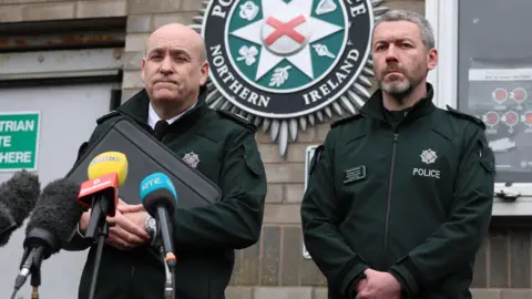 PA Media Assistant Chief Constable Ryan Henderson and District Commander Superintendent Brendan Green stand outside Lurgan police station. Henderson is stood in front of a collection of microphones with a neutral expression on his face. Green is on his left.