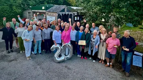 A group of volunteers are gathered out side the community ship cabin with signs that say 20 celebrating its 20th birthday. They are all smiling and looking at the camera with the cabin behind them amongst threes and bushes.