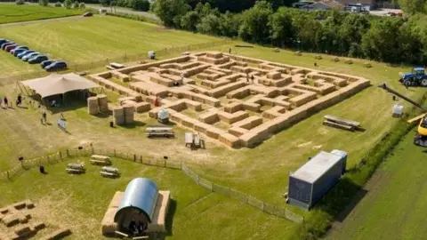 An overhead shot shows hundreds of straw bales laid out in an intricate maze in a giant field, with farm buildings dotted around the outside of it. A line of large leafy trees are on the horizon above the laid out bales.