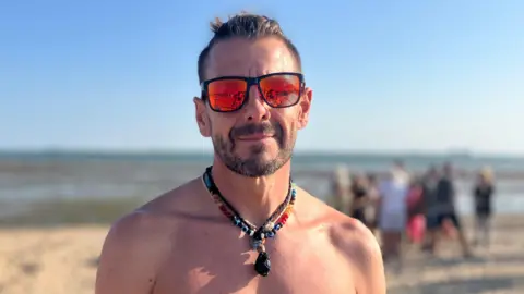 Henry Godfrey-Evans/BBC Matthew Dilley is bare-chested. He wears sports sunglasses and a beaded necklace. People can be seen gathered in a group behind him on the sands.