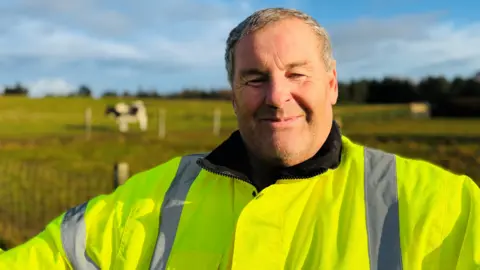 Mike Duxbury smiling at camera, he is wearing a bright yellow safety jacket, there is a cow in a field in the distance behind him.