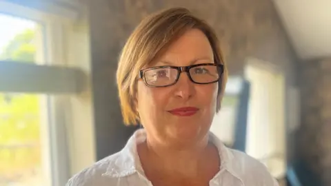 A woman with short, light brown hair, smiling at the camera. She is wearing glasses and a white shirt. She is pictured from the shoulders up. 