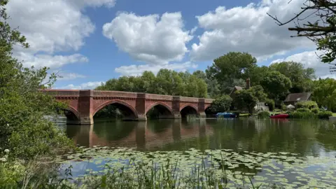 youngonce A brick arch bridge over a river on a sunny day. The sky overhead is blue with white clouds. Both banks of the river are lined with trees. There is a building on the far bank with a red and blue boats moored in the water.