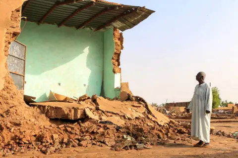 AFP An elderly man stands in front of the collapsed mudbrick wall of a house following floods in the Masawi area near Merowe in Sudan's Northern State on August 28, 2024.