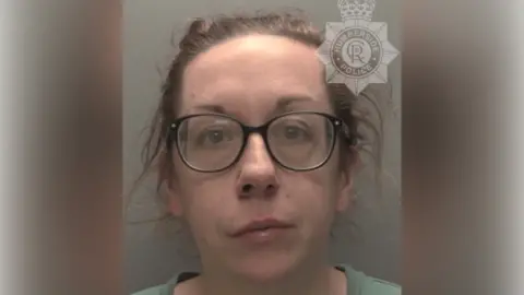 A police custody shot of Gemma Phoenix looking at the camera. She has brown hair tied back and is wearing a green t-shirt and black-rimmed glasses. The wall behind her is grey.