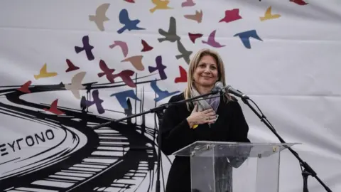 Getty Images The mother of a victim stands with her hand against her chest against a white backdrop decorated with flying birds