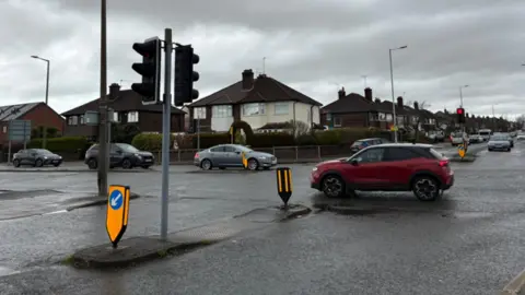 A number of cars are passing through a junction with traffic lights. Semi-detached houses can be seen.