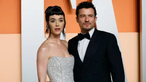 Reuters Katy Perry with Orlando Bloom at the Vanity Fair Oscars party. She is wearing a shoulderless silver dress, while he is wearing a traditional black and white tuxedo. 