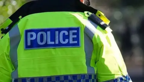 A picture of a police officer from the back. Their head is out of shot and they are wearing a bright jacket with the word POLICE on it.