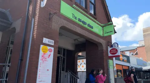 The front of the Co-Op on The Avenue. A two-storey red brick building, a green sign reading "The co-operative" sits above the entrance. Beneath it and to the right is a Post Office sign. A lane goes down the right hand side of the shop as we look at it.