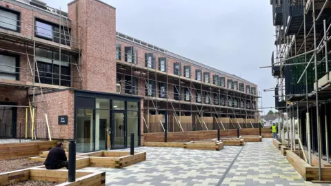 Local Democracy Reporting Service A man sitting on the edge of wooden plant beds which are outside modern three-storey flats which are shrouded in scaffolding. There is a grey and white paved area between two rows of flats.