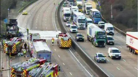 National Highways The M25, with traffic moving in four lanes on the other side of the road but the nearer side of the road is closed. There are a number of fire engines on the carriageway as well as a burnt-out lorry.