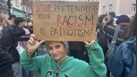 A women in a green hoody holds a sign that says 'Stop pretending your racism is patriotism'