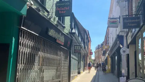Owen Sennitt/BBC Shutters have been lowered at a restaurant in Norwich city centre, with people walking by. The restaurant is painted black but has a sign saying "moorish falafel bar" in green and pink writing. People can be seen walking up the street in the distance.