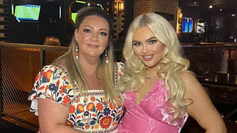 Breege Gormley Breege is wearing a white and multi-coloured dress with multi-coloured earrings. She has long dark brown hair and is standing beside her daughter, Jenny. Her daughter has long curly blonde hair and is wearing a pink dress. They are both standing in a bar. 