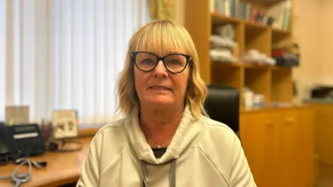 Una mujer rubia de cabello hasta los hombros mira a la cámara. Lleva gafas y una sudadera con capucha color beige. Detrás de ella hay un escritorio con un teléfono.
