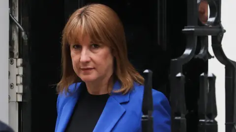 Reuters Chancellor Rachel Reeves wearing a dark blue jacket emerges from the doorway of 11 Downing Street
