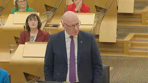 Scottish Parliament John Swinney, wearing a blue suit, stands to speak in the Scottish Parliament