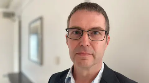 A man is looking into the camera. He is wearing black glasses and a jacket with shirt. He's standing in a large room.