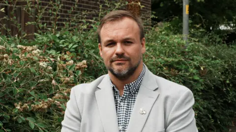 John Fairhall/BBC man in grey jacket and checked shirt - he has a beard and wears a lapel pin
