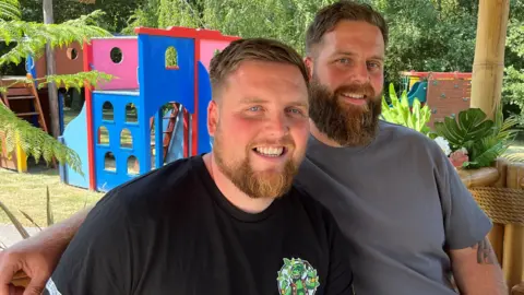 Two men both wearing t-shirts smiling at the camera. Behind them is a play area.