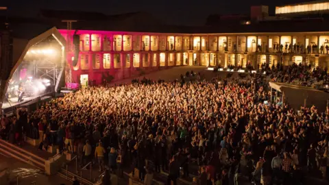 Getty Images The exterior of a bricked building showing a central courtyard filled with crowds of people who are watching a performance on a stage.